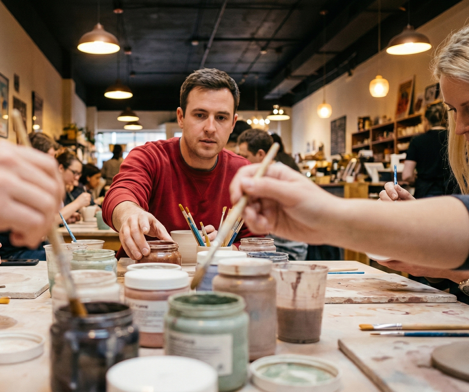 Grupo de personas pintando y esmaltando piezas de cerámica en un taller, mesa con botes de esmalte y pinceles, ambiente de aprendizaje y trabajo en equipo.